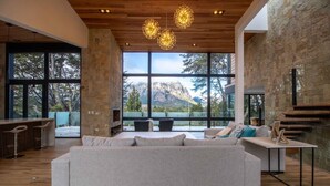 A spacious living area with a large sofa, a coffee table, and a bar area with stools. Large windows offer a view of mountains and trees. - New Modern Home, Golf, Polo, Few Minutes From Catedral Ski Resort.  (San Carlos de Bariloche)