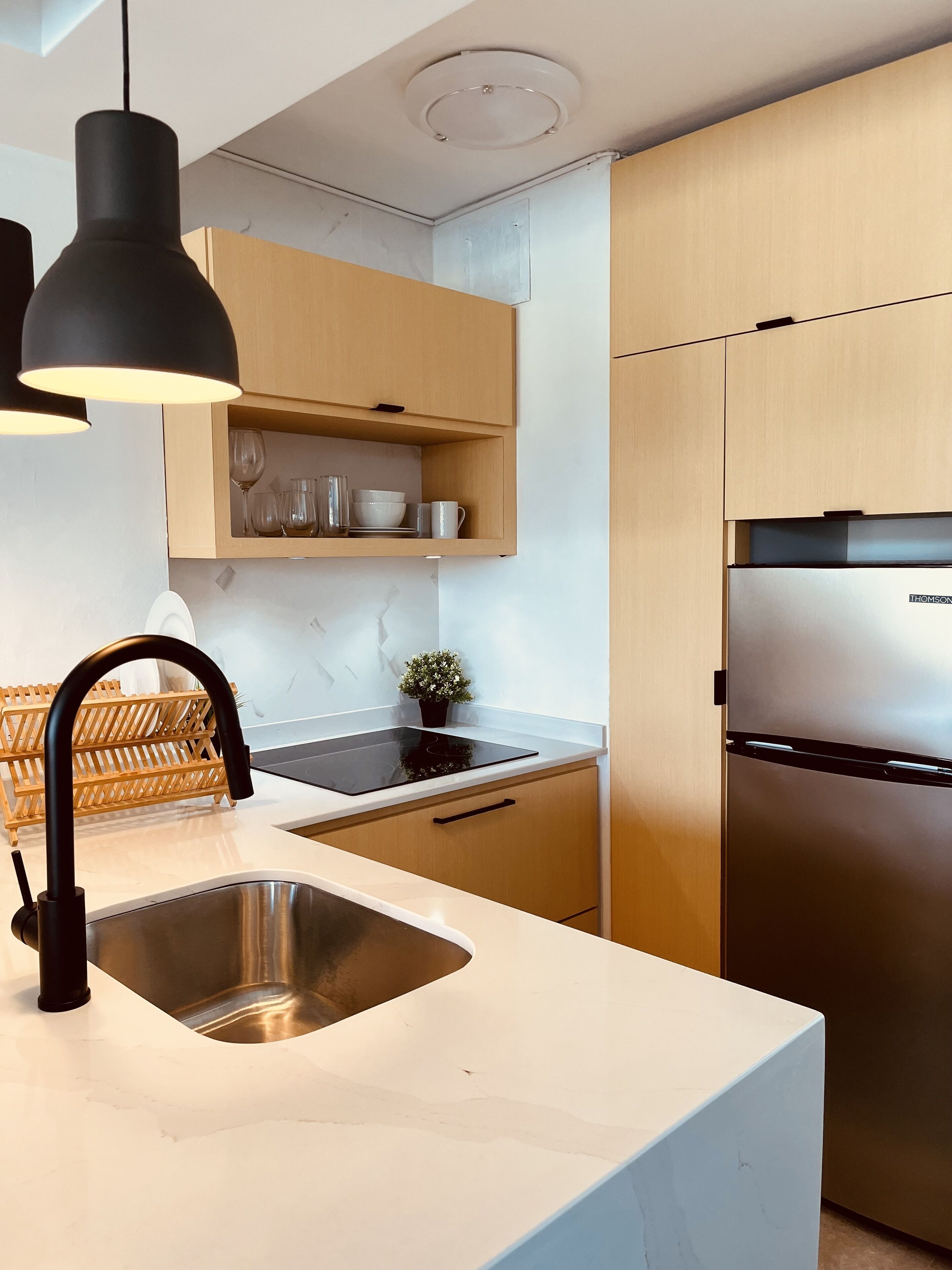 Une cuisine moderne dotée d’un évier en acier inoxydable, de luminaires suspendus noirs, de meubles en bois et d’un réfrigérateur encastré.