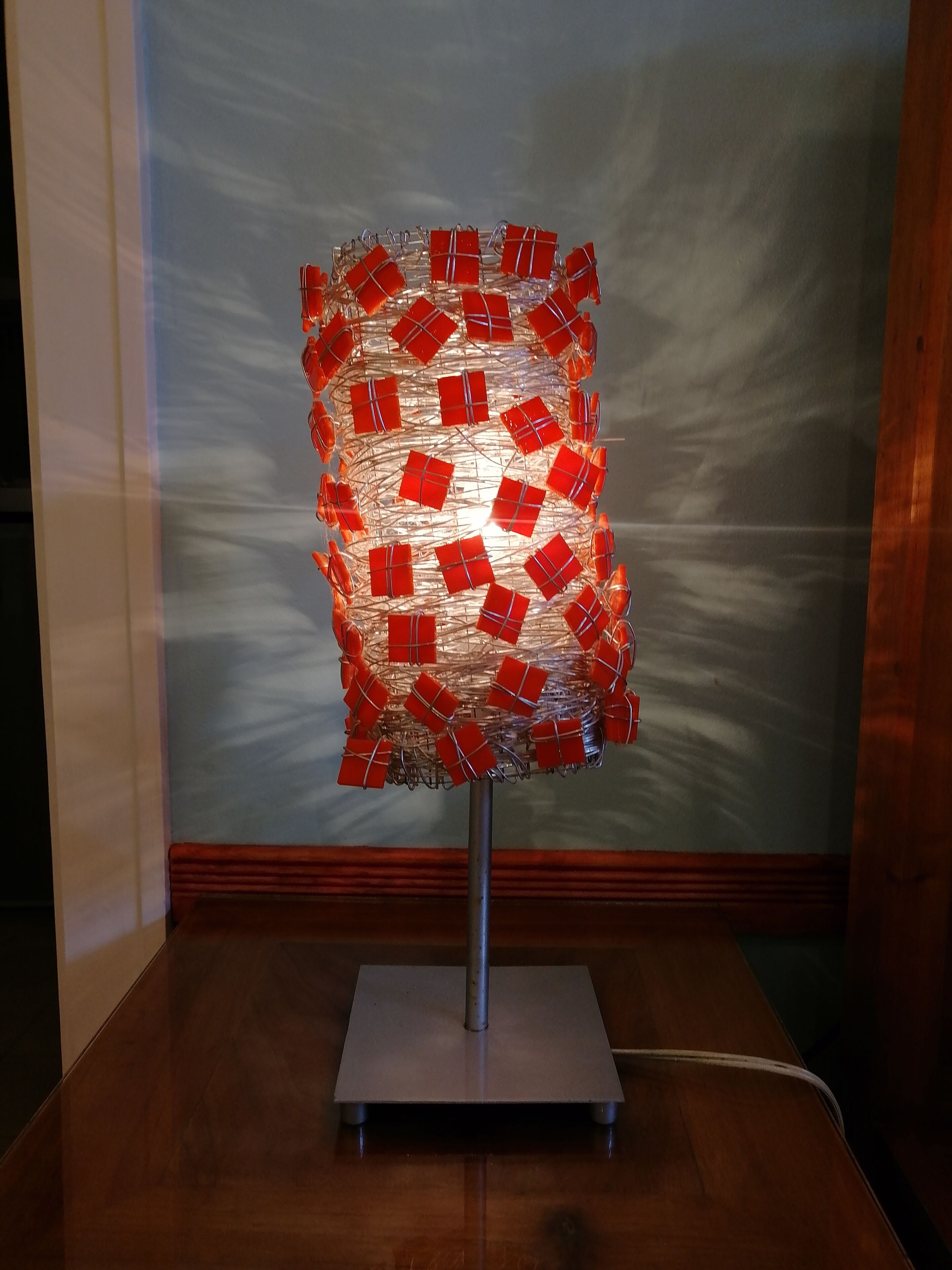 A sculpture with red and clear elements on a metal stand in a room with wooden flooring and a plain wall.