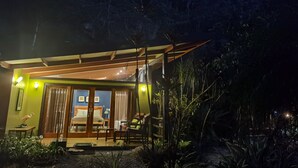 Een gezellige, goed verlichte kamer met een bed, een kleine tafel en een stoel, omgeven door weelderige begroeiing en uitzicht op een beboste heuvel.