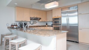 Una cocina moderna con armarios de madera clara, un refrigerador de acero inoxidable, una encimera de granito y una lámpara colgante.