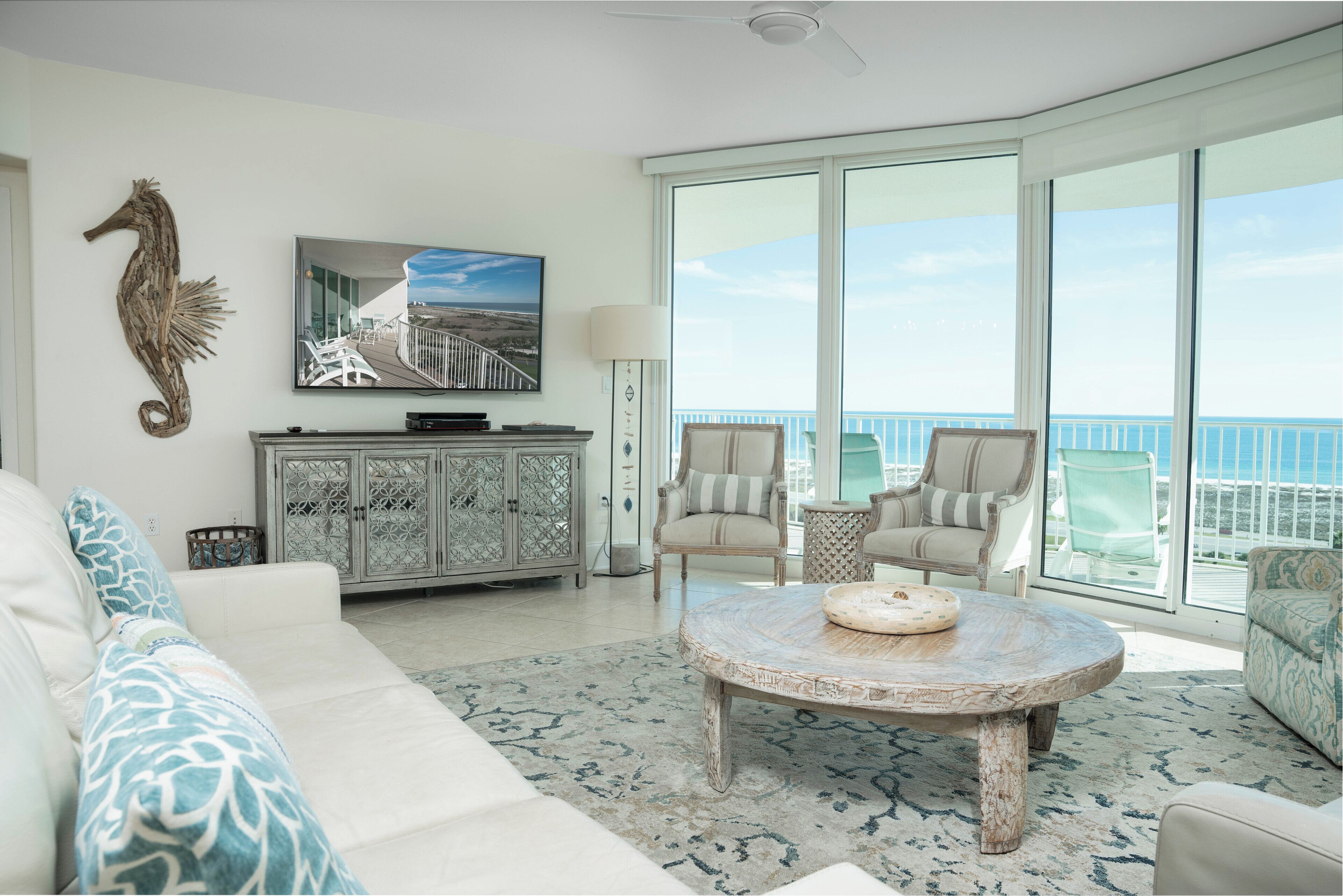 Uma sala de estar espaçosa com uma grande janela que oferece vista para o oceano, um sofá branco, duas poltronas, uma mesa de centro e um armário com objetos decorativos.