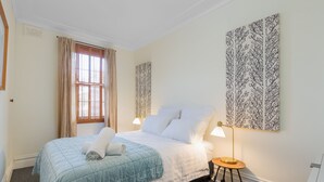 Un lit bien fait, recouvert d’une courtepointe bleue, une lampe de chevet et un mur orné d’une œuvre d’art à motifs de feuilles.