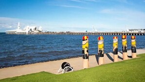 A waterfront view with a pier extending into the water, a white building with a tower, and a row of bollards with yellow and red markings. - ⭐BROOKS⭐BFast,WiFi,Nflx⭐UberEats, Walk2Shop, (Norlane)