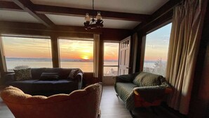 A living room with a view of the sunset over the water, a sofa, a chair, and a chandelier. - Windsurfers Cove, shore front cottage (Tiverton)