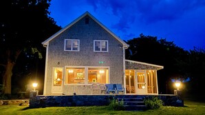 A two-story house with a stone facade, large windows, and a porch with chairs. - Windsurfers Cove, shore front cottage (Tiverton)