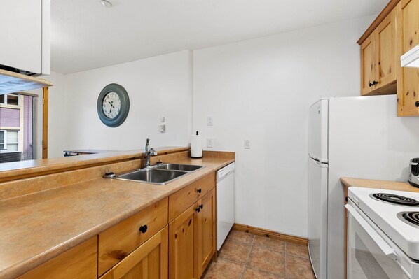 Cucina compatta con mobili in legno, lavandino in acciaio inossidabile, lavastoviglie bianca, frigorifero bianco e piano cottura.