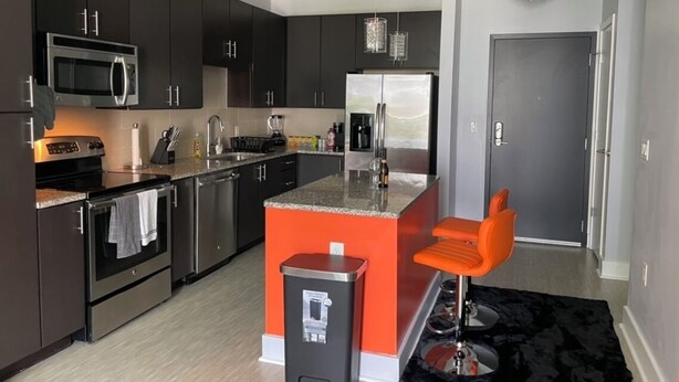 A modern kitchen with dark cabinets, stainless steel appliances, and a central island with orange bar stools.