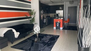 A modern living room with a black sofa, a glass coffee table, and a black rug. There is a kitchen in the background with dark cabinets and a refrigerator.