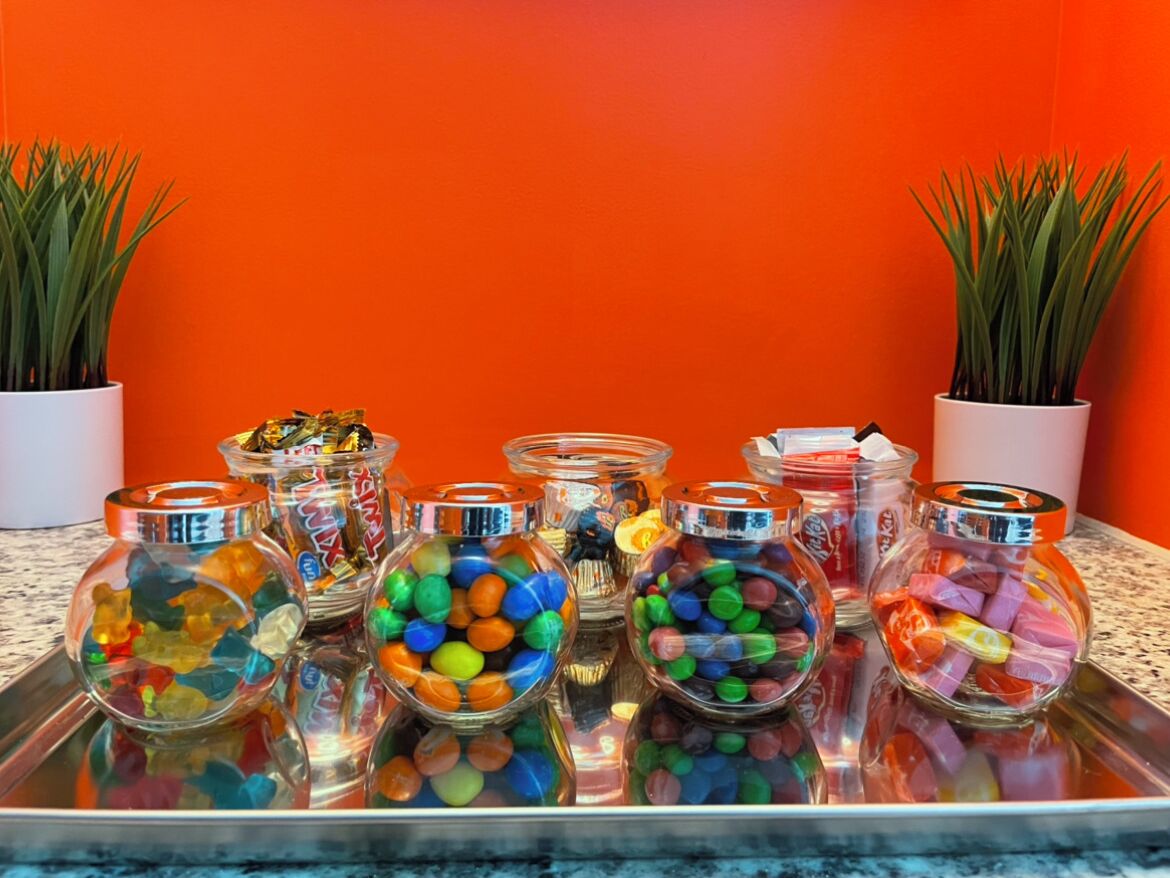 A tray with glass jars containing colorful candies and a jar with wrapped candies, placed on a marble countertop with orange walls and potted plants.