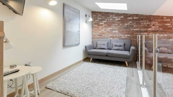 A modern living room with a grey sofa, a small white table with a remote control, a grey rug, and a brick wall accent.