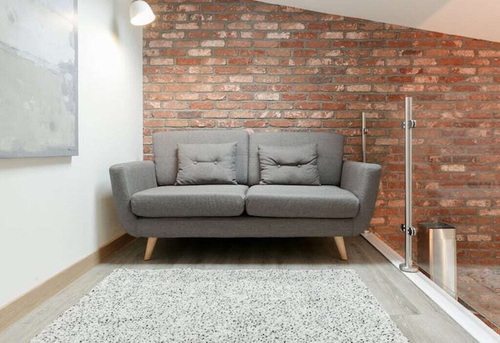 Ein modernes Wohnzimmer mit grauem Sofa, einem strukturierten Teppich und einer Ziegelwand.
