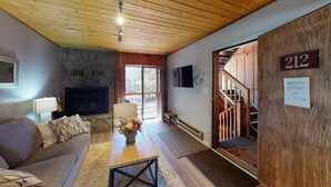 A cozy room with a stone fireplace, wooden ceiling, a sofa, a coffee table with flowers, and a staircase leading to another level. - Fireplace | WiFi | Close to Lifts | Windchimes 212 (Angel Fire)