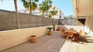 A patio area with a brick floor, wooden furniture, potted plants, and a tall fence. - Puerto Romano II 88 by homeprive (Denia)