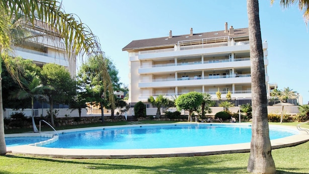 A swimming pool surrounded by a well-maintained lawn, with a multi-story building in the background. - Puerto Romano II 88 by homeprive (Denia)