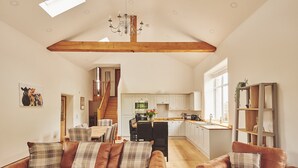 A spacious living room with a high ceiling, a wooden beam, and a chandelier. There is a kitchen in the background with white cabinets and a window. - Plum Cottage -  Luxury Cottages (Appleby)