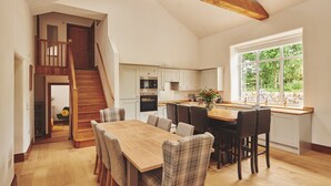 A spacious kitchen with a large island, wooden flooring, and a view of greenery through a large window. - Plum Cottage -  Luxury Cottages (Appleby)