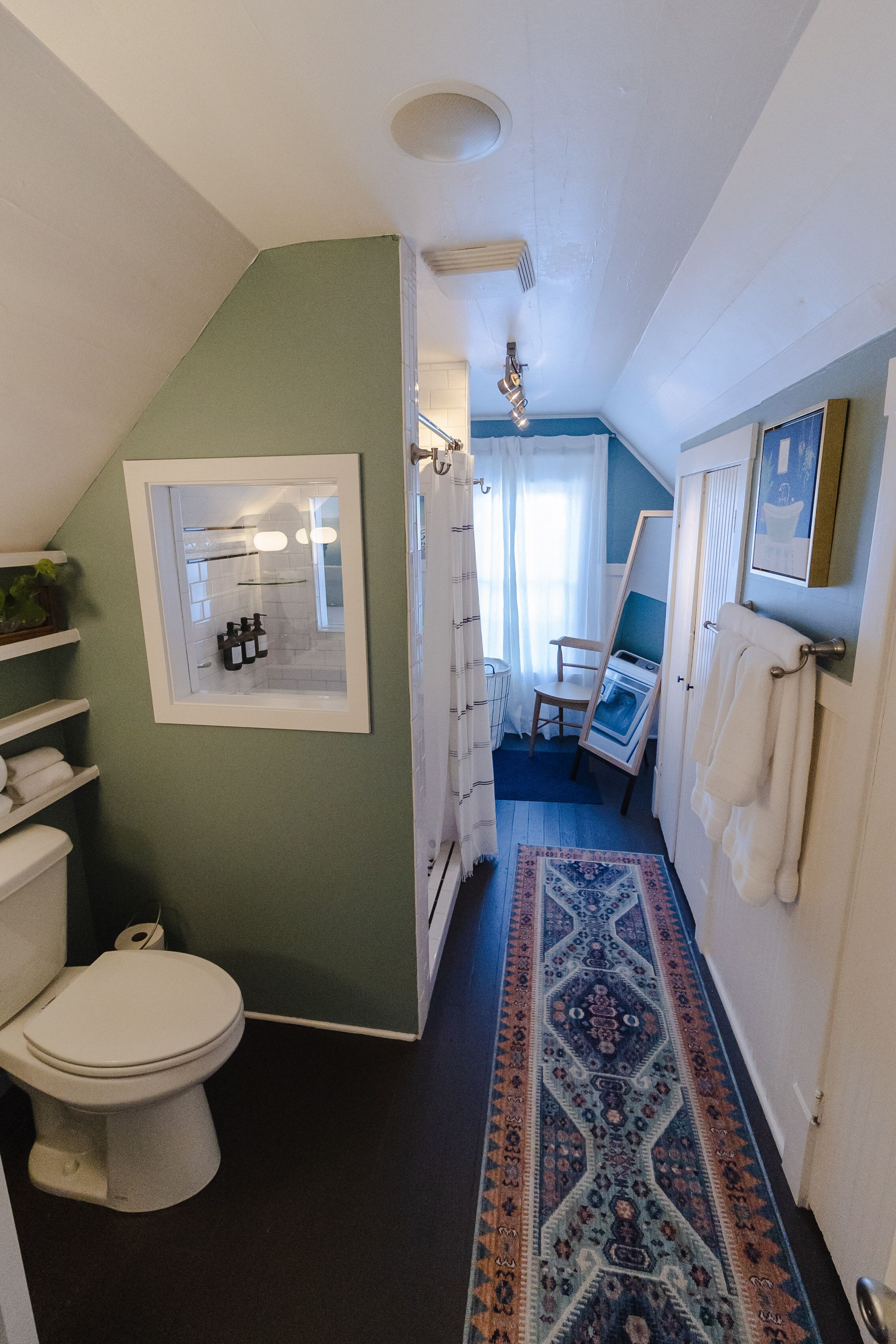 A narrow hallway with a patterned rug, a toilet, a framed mirror, and a door leading to a room with a chair and a painting.
