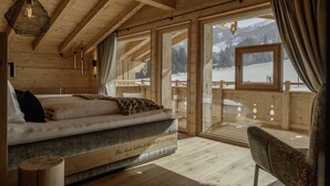 A wooden cabin bedroom with a bed, a chair, and a large window overlooking a snowy landscape. - Stylish chalet with private sauna, terrace and outdoor shower (Reith im Alpbachtal)