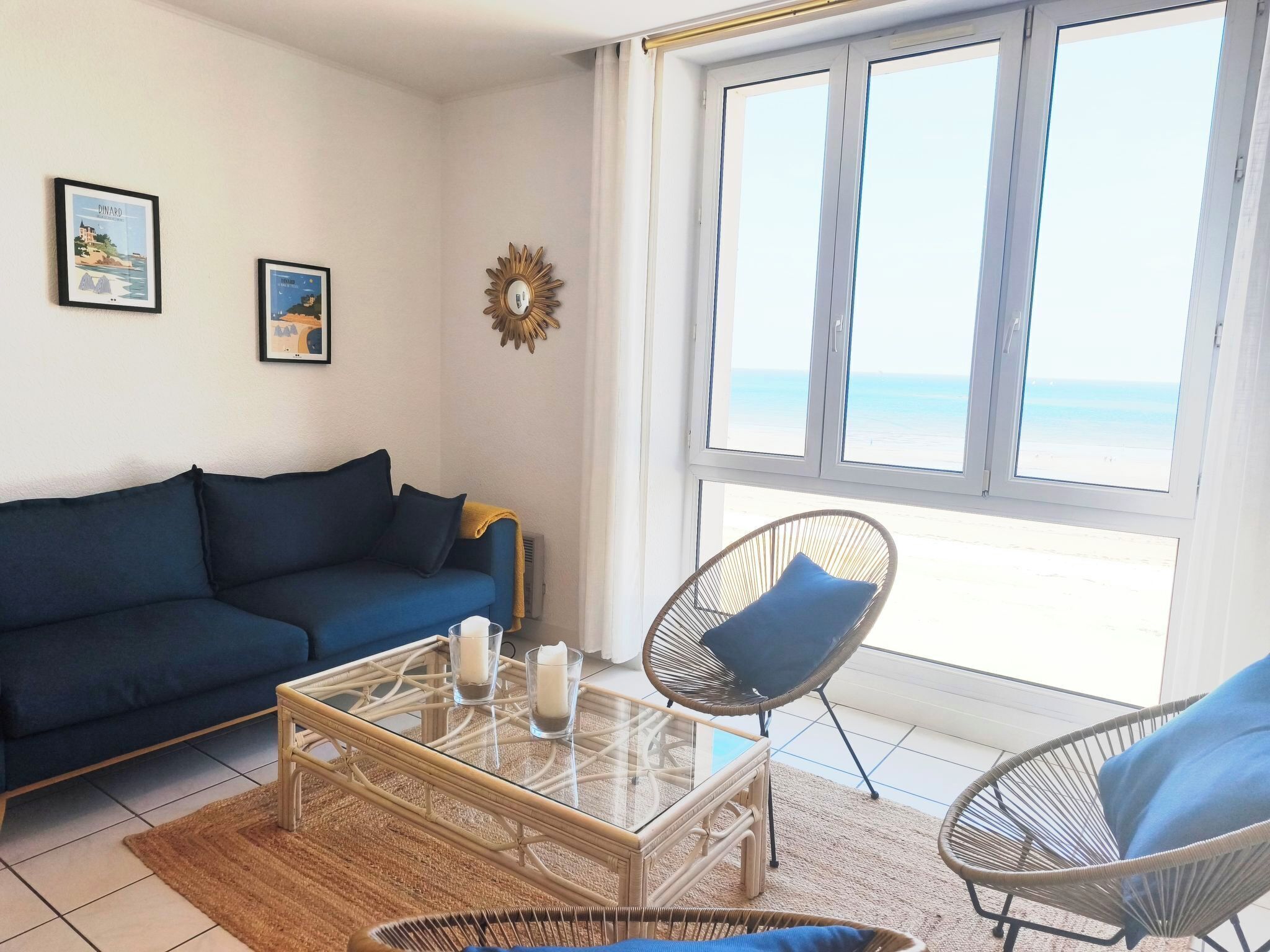 Un saló amb un sofà blau, una taula de café de vidre amb espelmes, cadires de vimet i una finestra gran amb vista a la platja.