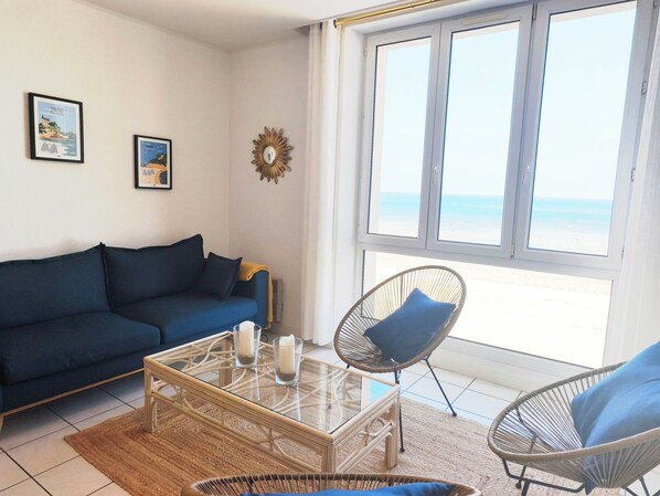A living room with a blue sofa, a glass coffee table with candles, wicker chairs, and a large window with a view of the beach.