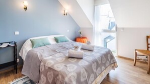 A bedroom with a sloped ceiling, a bed with patterned bedding, two wall-mounted lights, a small table with a vase, and a wooden chair. - Kerisper by Interhome (La Trinité Sur Mer)