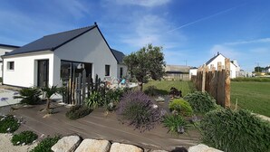 Une maison moderne dotée d’une grande porte vitrée, entourée d’un jardin bien entretenu où poussent différentes espèces de plantes, et agrémentée d’une terrasse en bois.