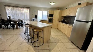 A modern kitchen with a central island, bar stools, wooden cabinets, a stainless steel refrigerator, and a dining area with a table and chairs. - Sandstone Point (SEQ) Bribie Holidays Family Home, pet friendly  (Sandstone Point)