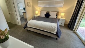 A bedroom with a bed, two bedside tables, a ceiling fan, a window with blinds, and a door leading to a bathroom. - Sandstone Point (SEQ) Bribie Holidays Family Home, pet friendly  (Sandstone Point)