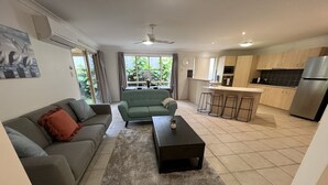 A modern living room with a grey sofa, a green sofa, a coffee table, and a kitchen area with a stainless steel refrigerator and tiled flooring. - Sandstone Point (SEQ) Bribie Holidays Family Home, pet friendly  (Sandstone Point)