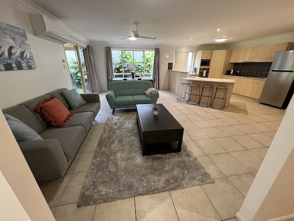 A modern living room with a grey sofa, a green sofa, a coffee table, and a kitchen area with a stainless steel refrigerator and tiled flooring. - Sandstone Point (SEQ) Bribie Holidays Family Home, pet friendly (Sandstone Point)