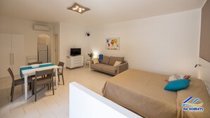 A compact hotel room with a bed, a sofa, a dining area with a table and chairs, a TV, and a small side table with a lamp.