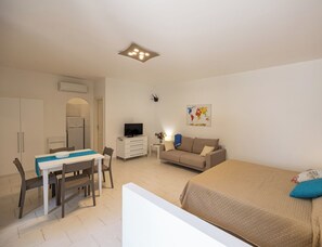 Quarto de hotel compacto com cama, sofá, área de refeições com mesa e cadeiras, televisão e pequena mesinha de cabeceira com abajur.