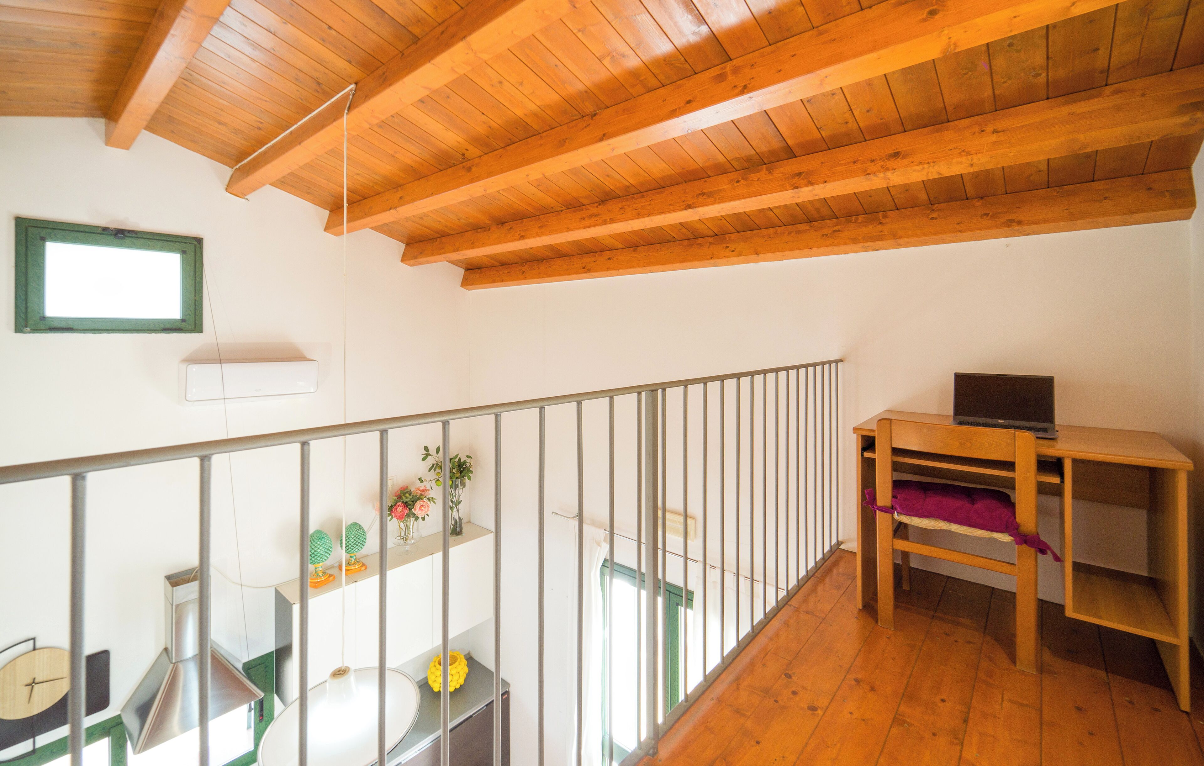 Techo de madera con lámpara colgante, baranda metálica, escritorio con una computadora portátil y ventana de marco verde.