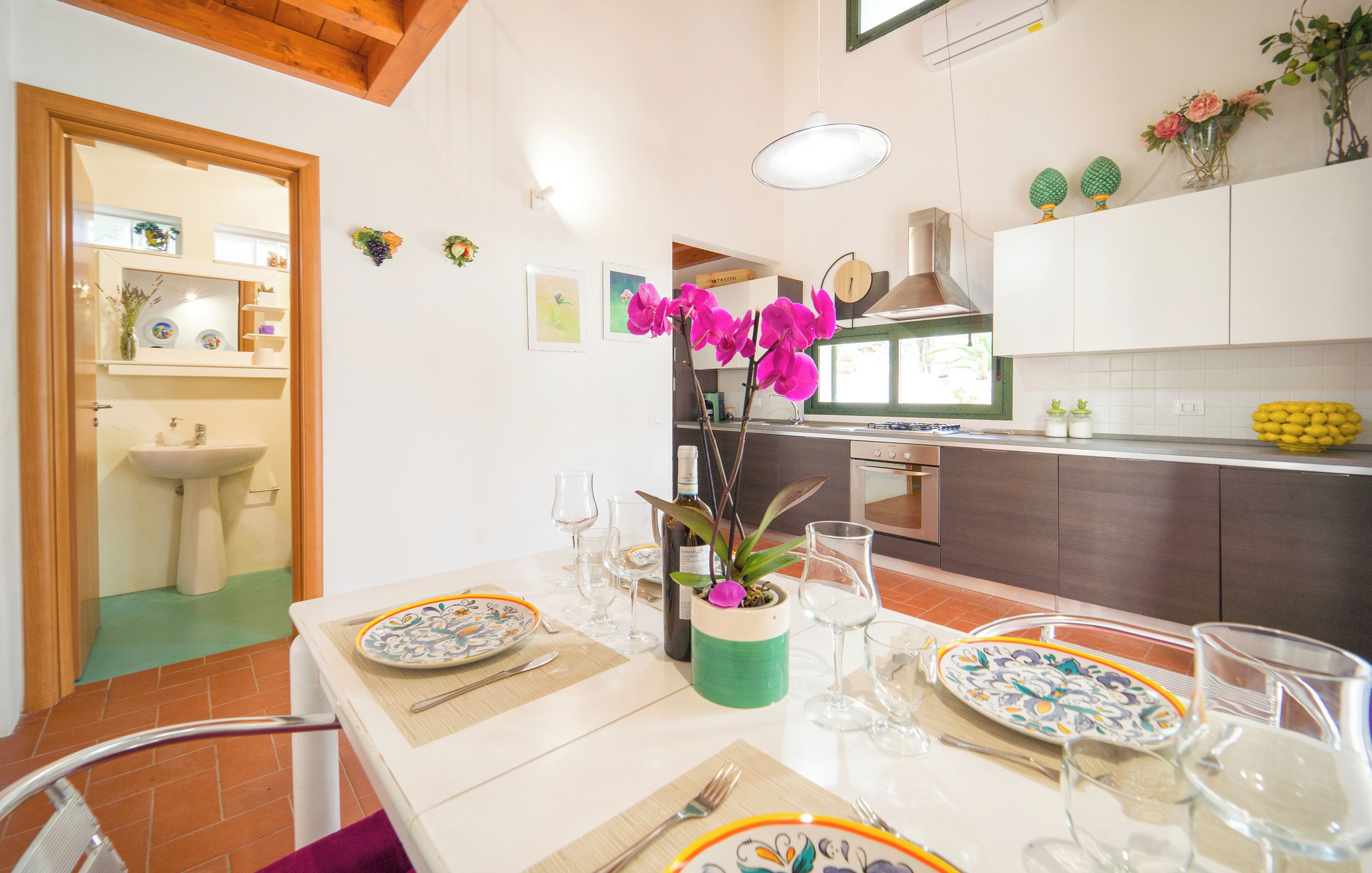 Un coin repas avec une table dressée pour deux personnes, une cuisine à l’arrière-plan et une salle de bain visible à travers une porte ouverte.