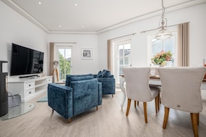 Ein modernes Wohnzimmer mit einem Flachbildfernseher, blauen Sofas, einem Esstisch mit Stühlen und einem großen Fenster.