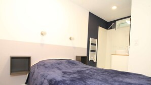 Une chambre à coucher comprenant un lit, une table de chevet, une étagère fixée au mur, et une salle de bain visible à travers une porte ouverte.