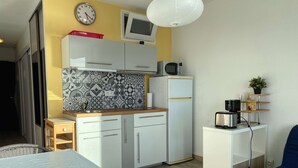 Une cuisine compacte avec des meubles blancs, un four à micro-ondes et un réfrigérateur. On y trouve une petite table avec une chaise, une table basse en verre et un canapé bleu.
