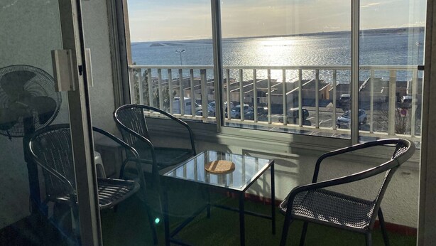 Un balcon avec une table et des chaises en verre, donnant sur une étendue d’eau et un parking.