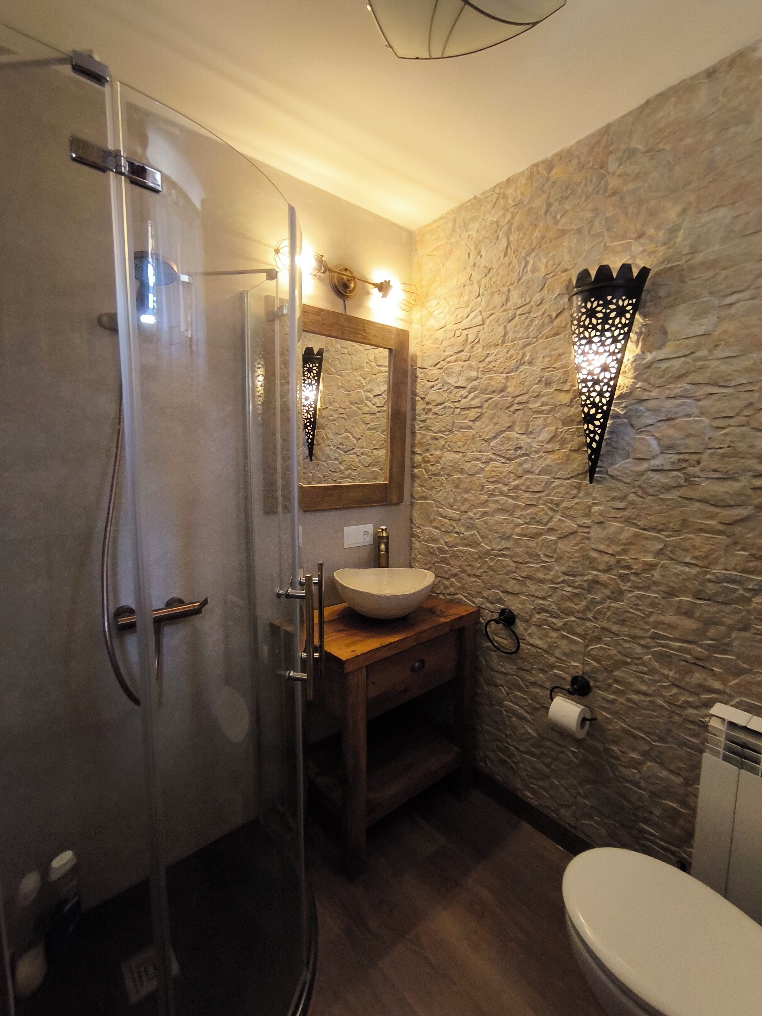 Une salle de bain dotée d’une douche à paroi de verre, d’un meuble-lavabo en bois avec un évier, d’un miroir et d’un mur à l’aspect pierre.