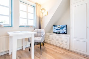 Quarto com escrivaninha branca, poltrona cinza, televisão e janela com cortinas.