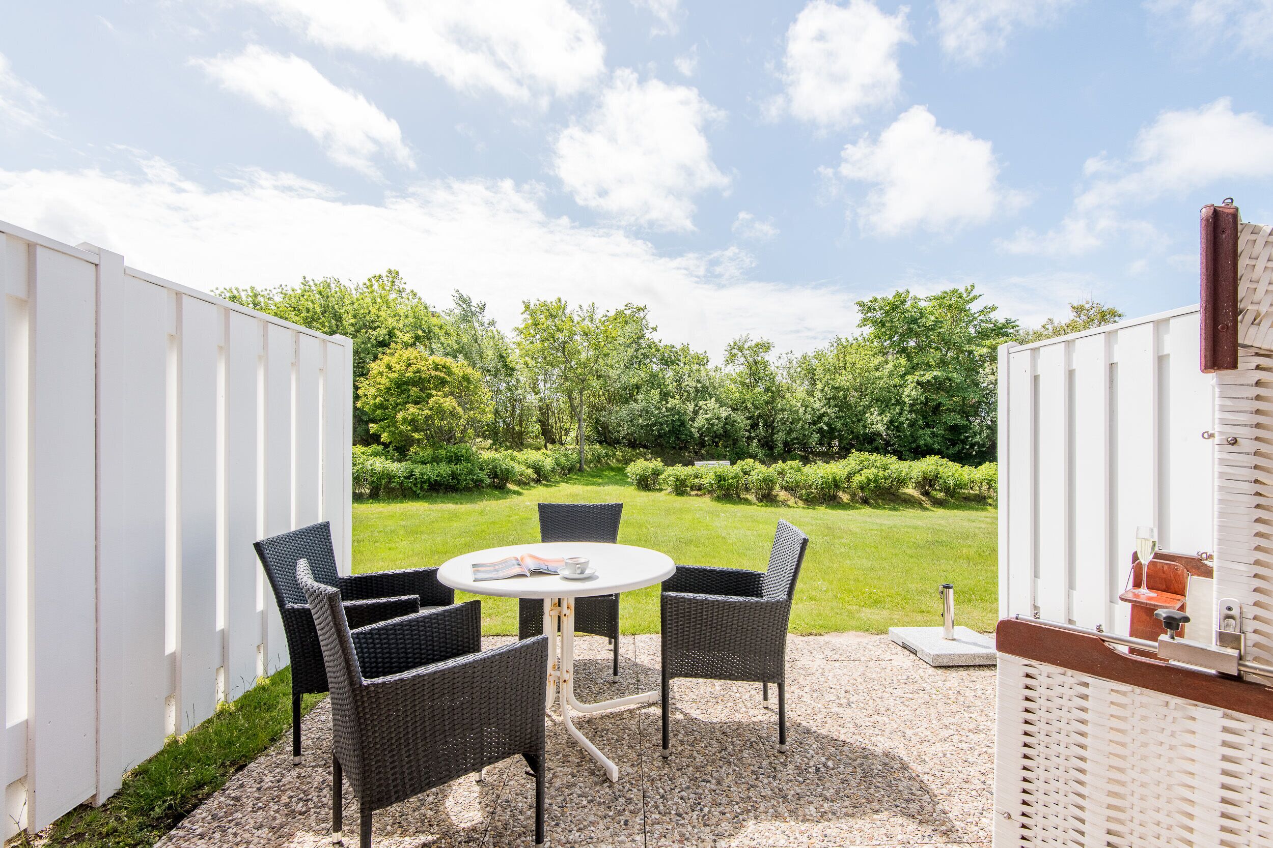 Un espace patio avec une table ronde et quatre chaises, entouré d’une clôture blanche, donnant sur une pelouse bien entretenue et des arbres.