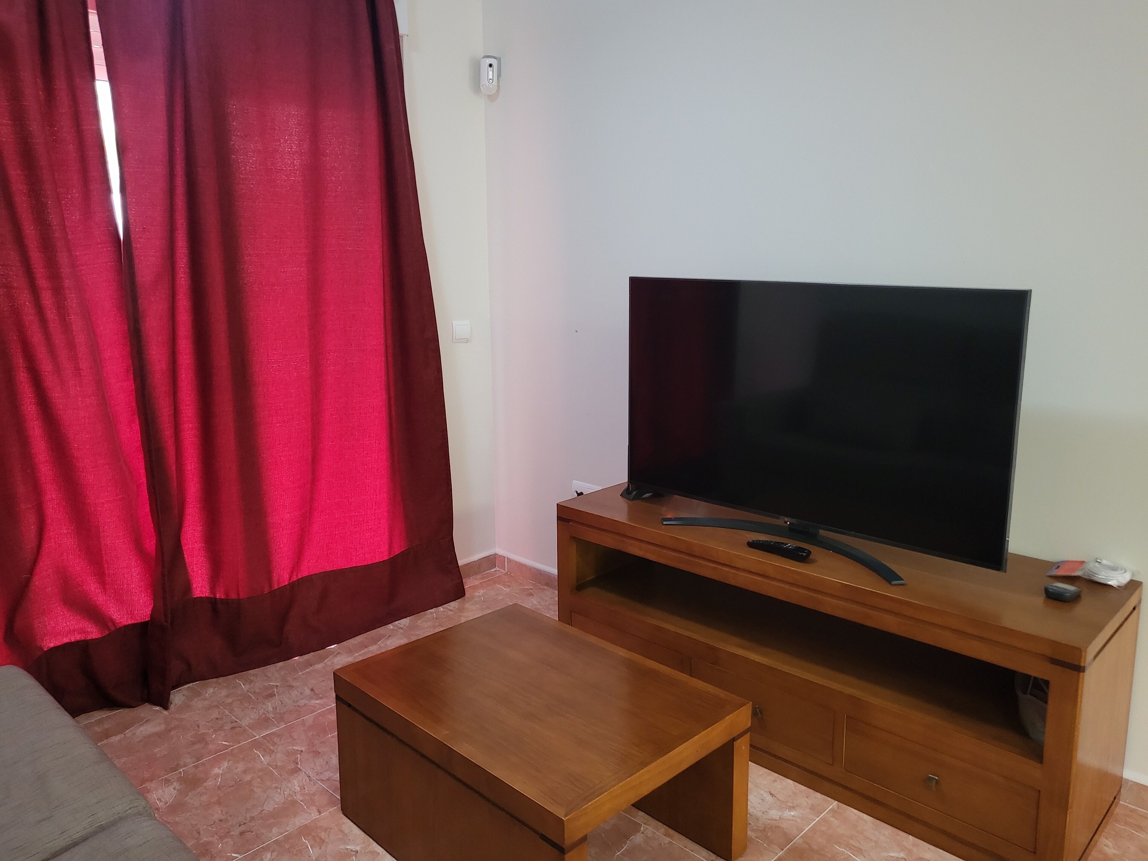 Ein Wohnzimmer mit einem Flachbildfernseher auf einem Holzständer, einem kleinen Holztisch und einem roten Vorhang.