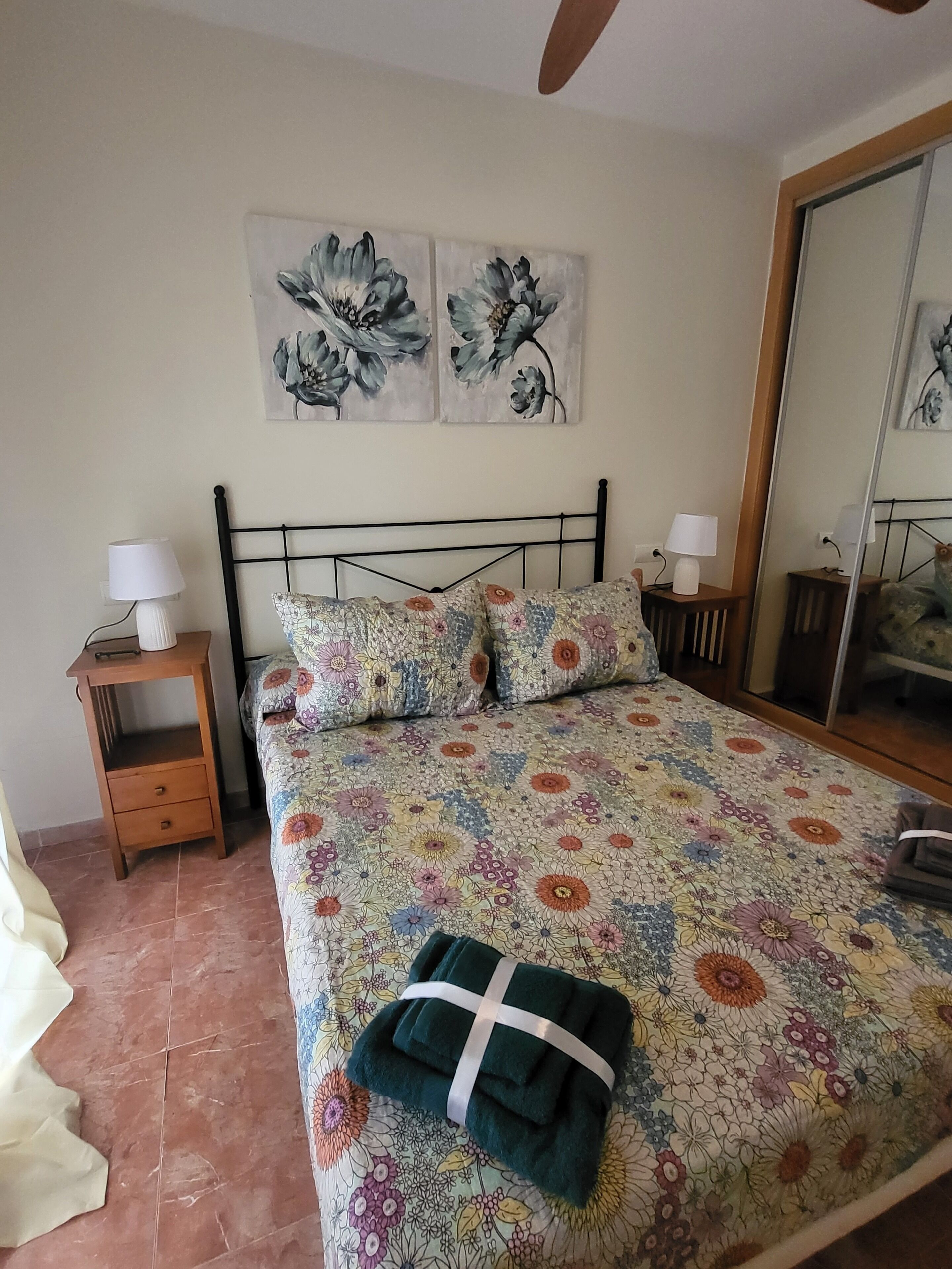 A bedroom with a floral bedspread, two wall-mounted paintings, a wooden nightstand with a lamp, and a mirror.