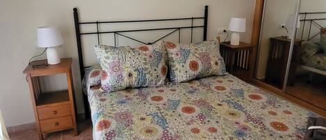 A bedroom with a floral bedspread, two wall-mounted paintings, a wooden nightstand with a lamp, and a mirror.