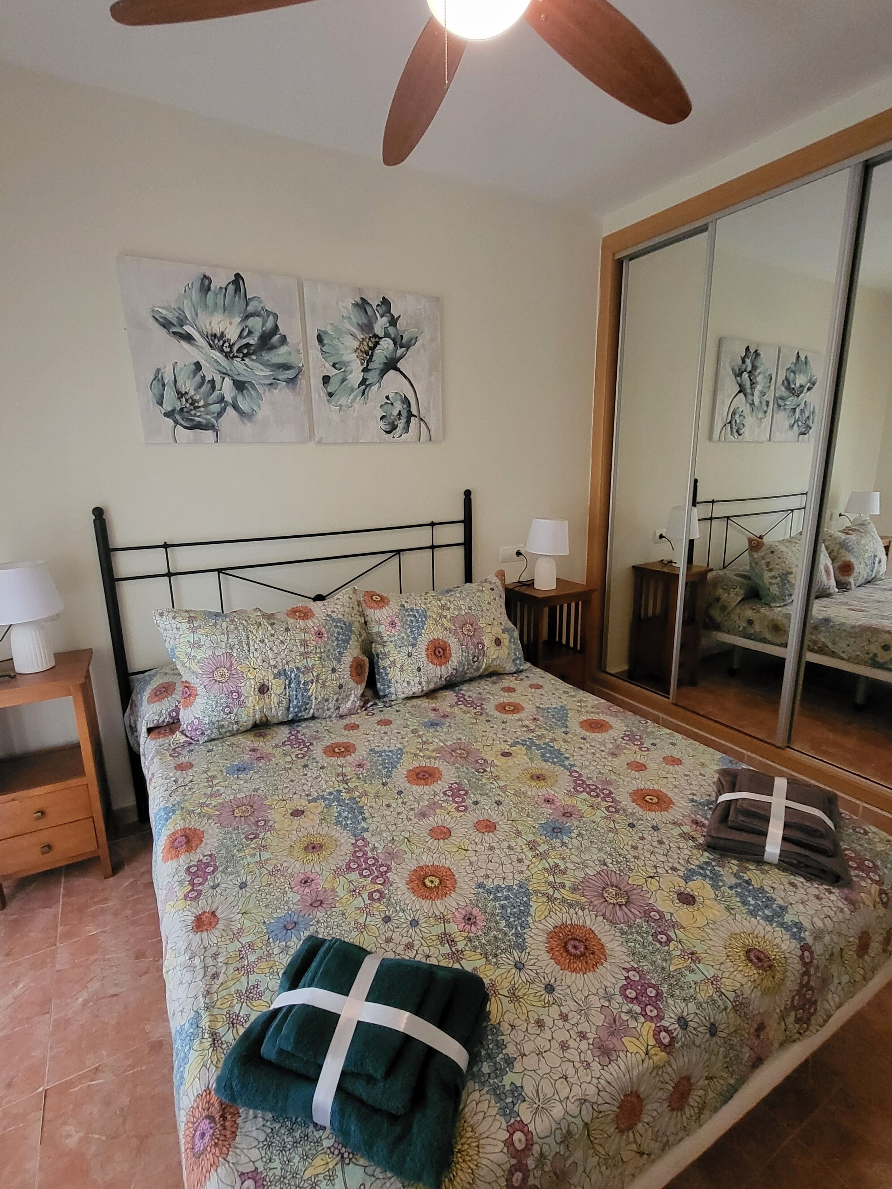 Une chambre à coucher comprenant un lit, une table de chevet, une lampe, un miroir et une œuvre d’art aux motifs floraux accrochée au mur.