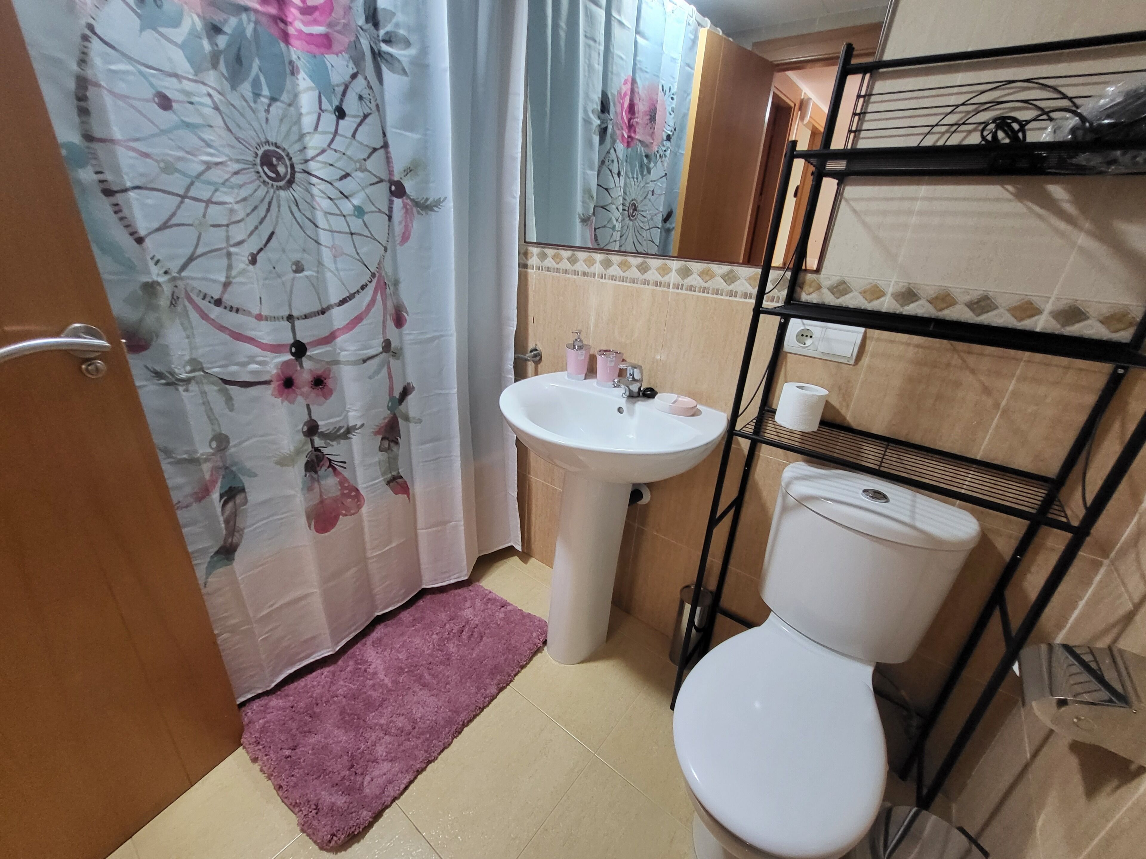 Une salle de bain avec un lavabo blanc, des toilettes, un rideau de douche à motifs floraux, un tapis rose et une étagère en métal contenant des articles de toilette.