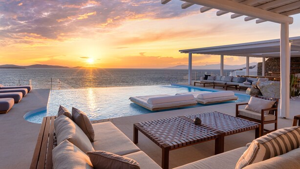 A poolside area with lounge chairs and a table, overlooking a body of water during sunset. - Villa Santa Esmeralda Mykonos (Mykonos)