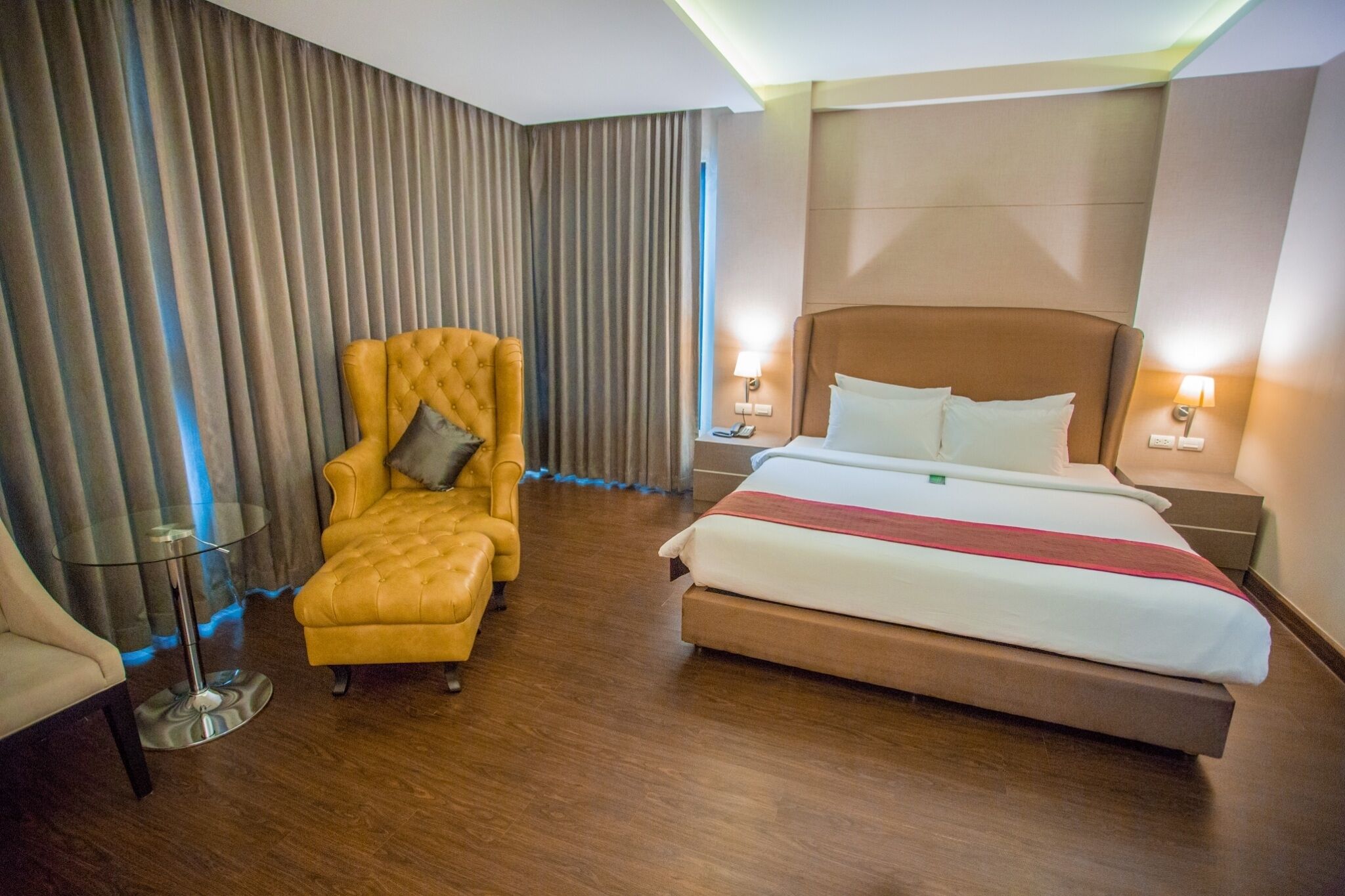 A hotel room with a large bed, a yellow armchair, a glass table, and a view of the city through the window.