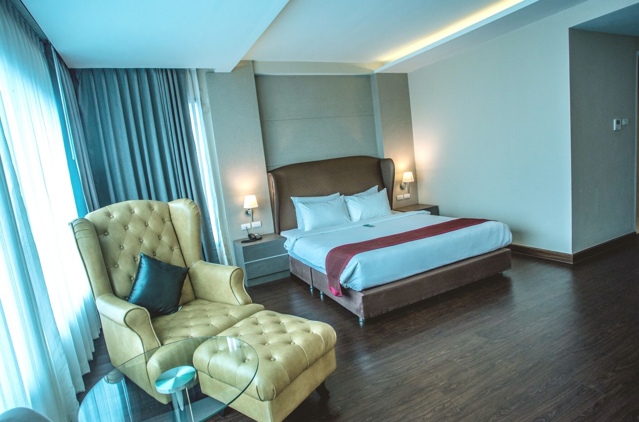 A hotel room with a large bed, a tufted armchair, a glass side table, and a nightstand with lamps.